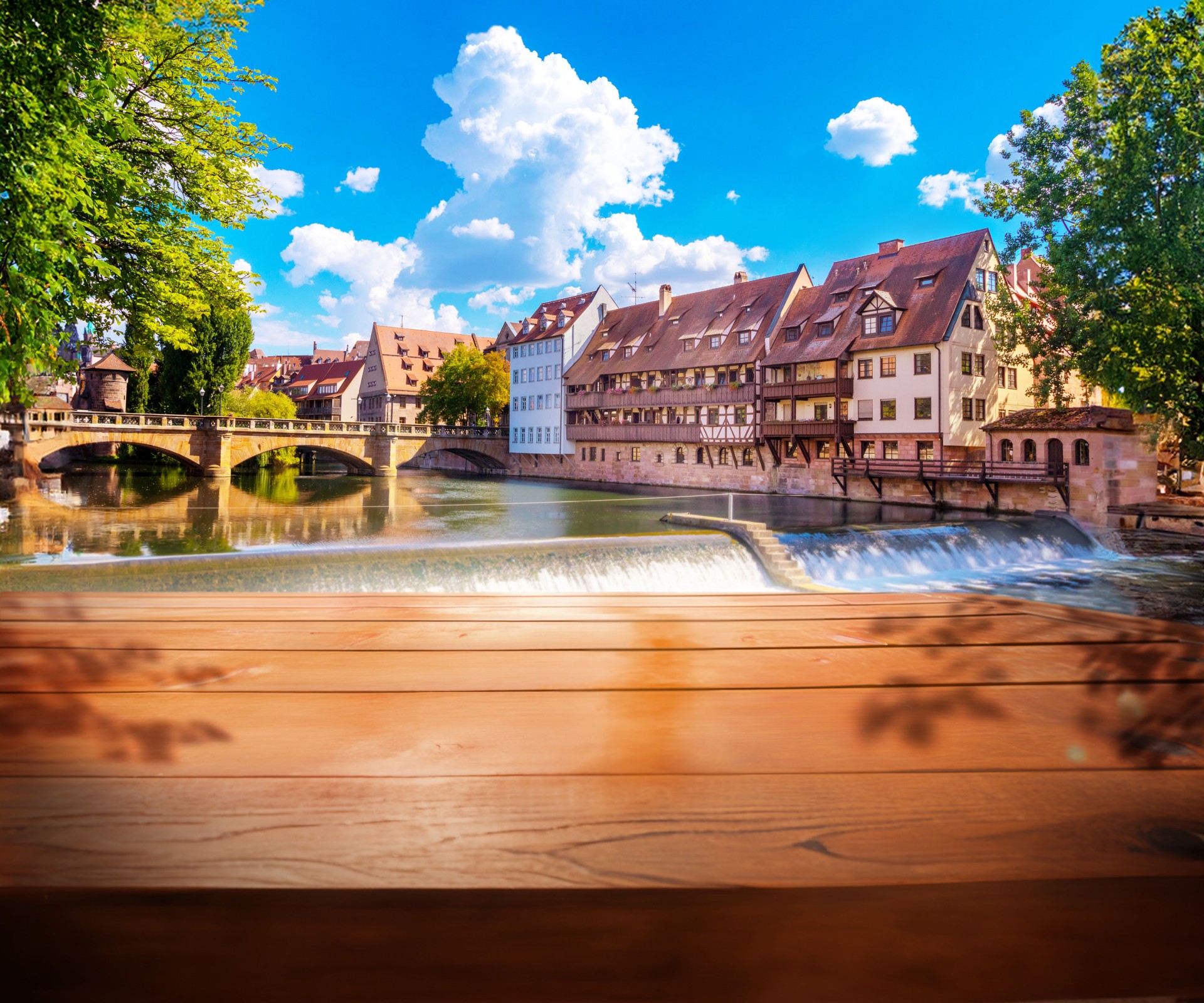Historic timber-framed houses stand beside a river, over which a bridge runs. In the foreground a wooden plank is visible.