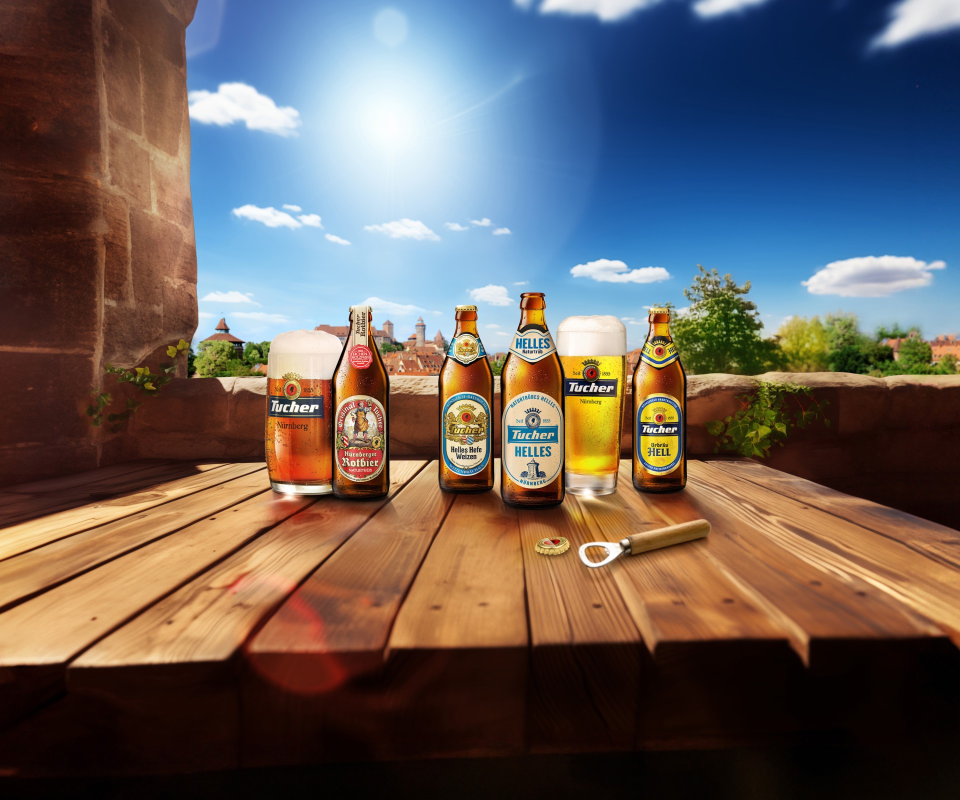Five bottles and a glass of beer sit on a wooden table in front of a sunny landscape.