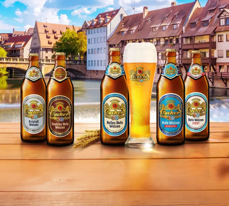 Five beer bottles and a full glass stand before a riverside scene with houses.
