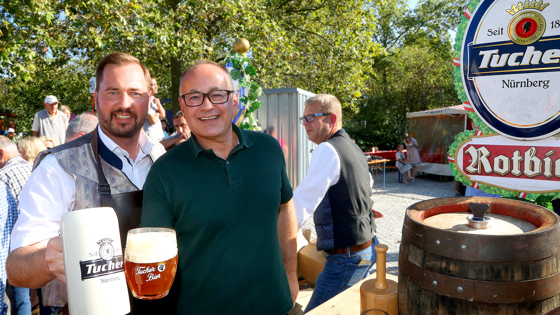 Two men smile and hold drinks at a sunny festival.
