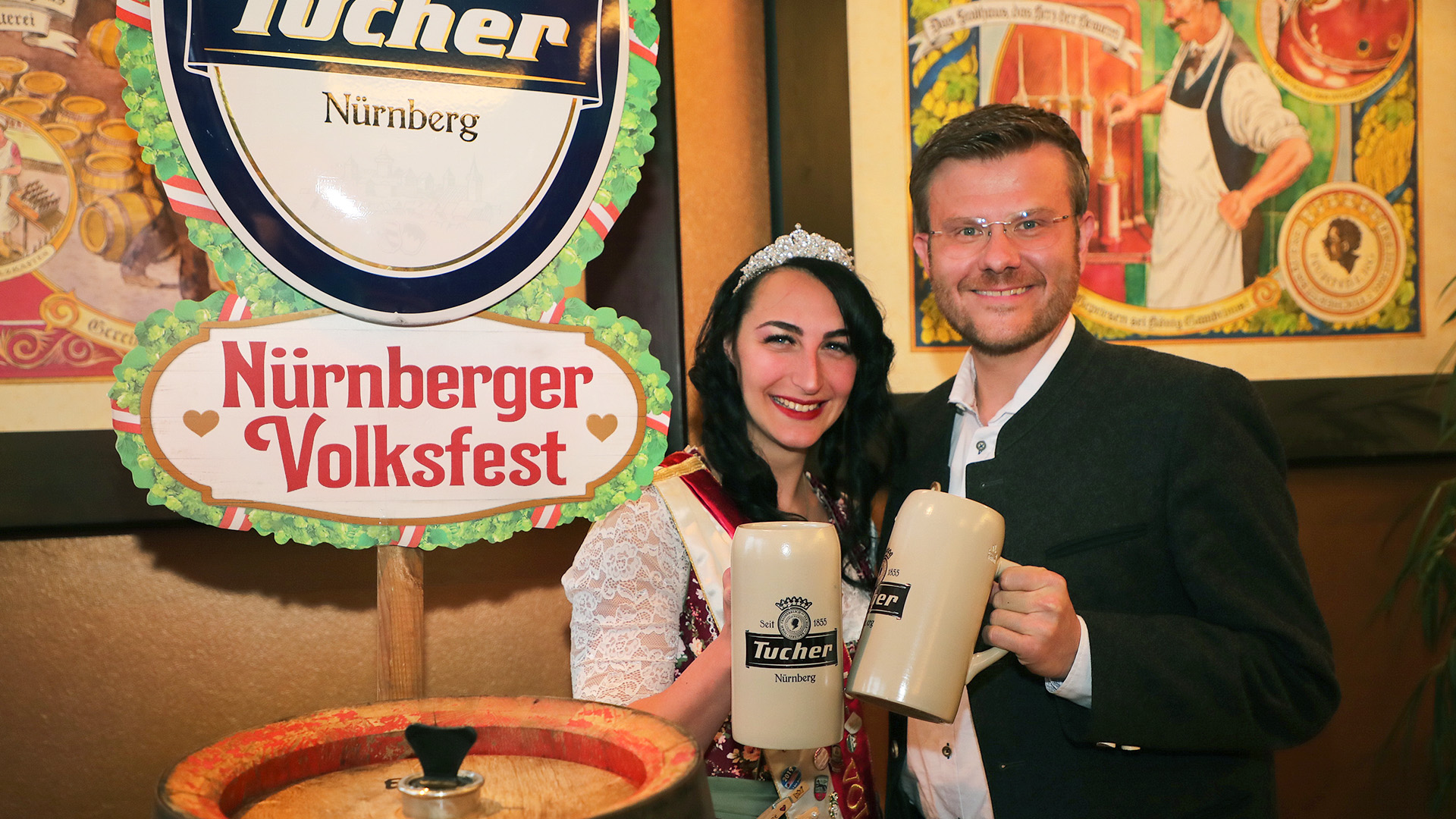 Two people smile and hold large beer mugs, surrounded by festival decorations.