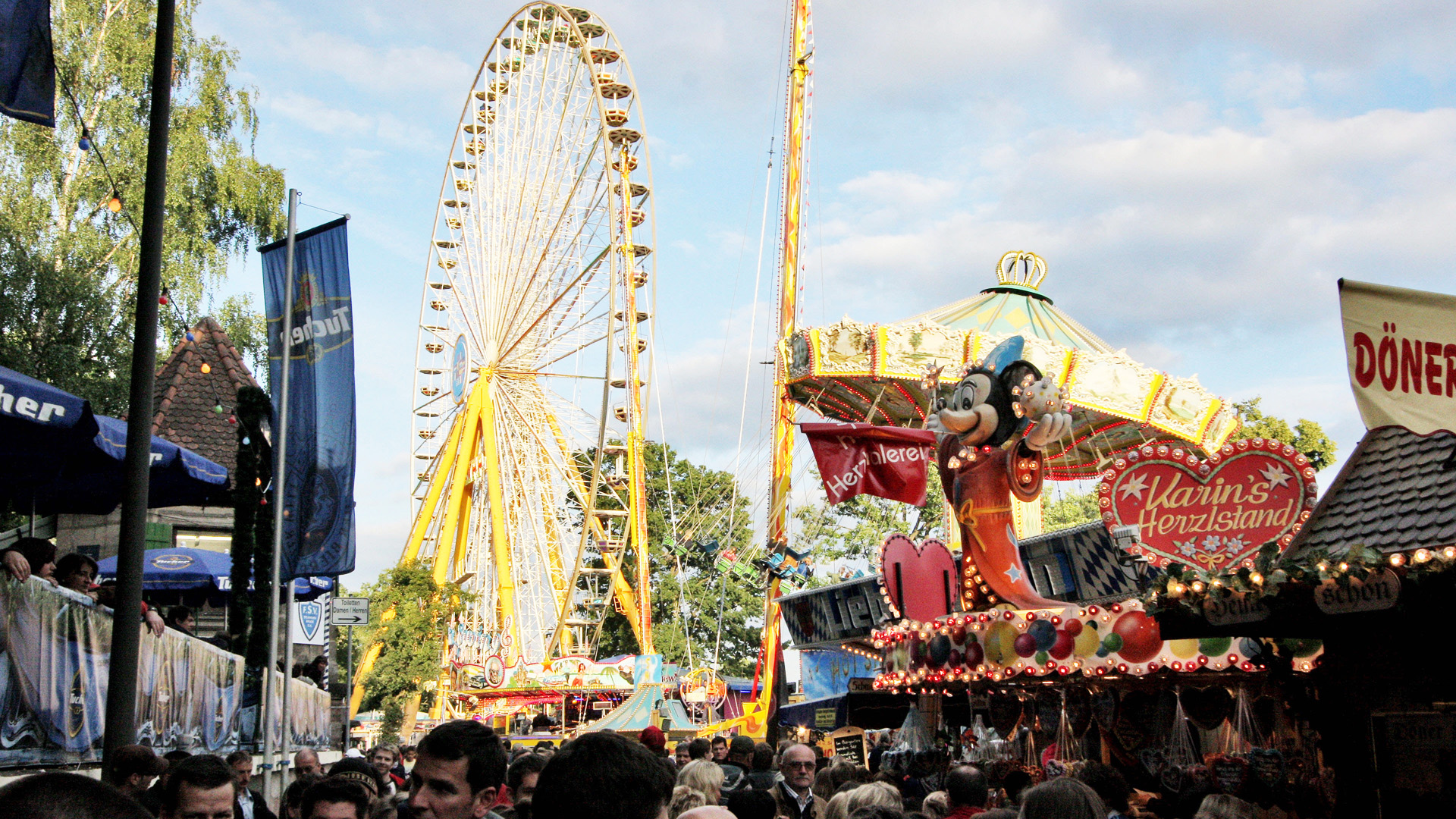 A bustling fair with a large Ferris wheel and various stalls in daylight.