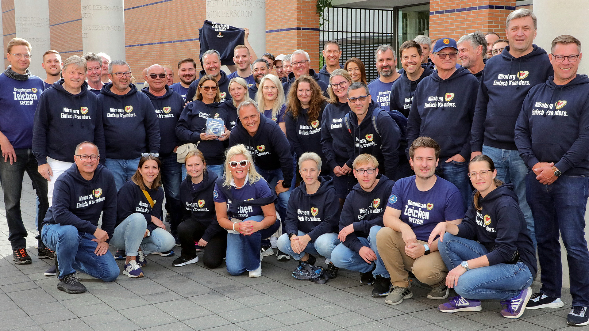 A group of people in blue hooded pullovers stands smiling in front of a building.