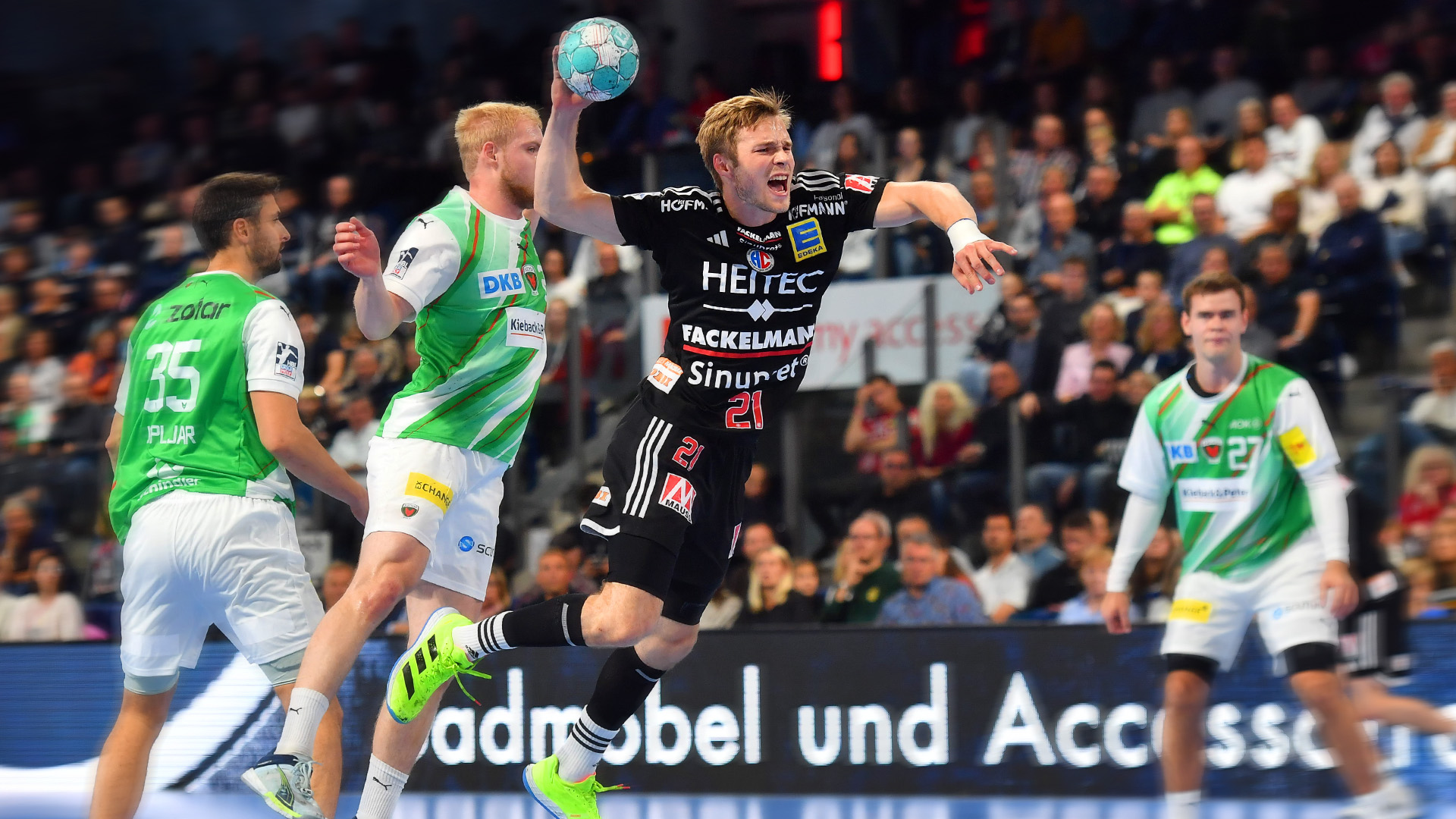 Three handball players contend for the ball during a match in a large hall. The player in the centre jumps and throws the ball.