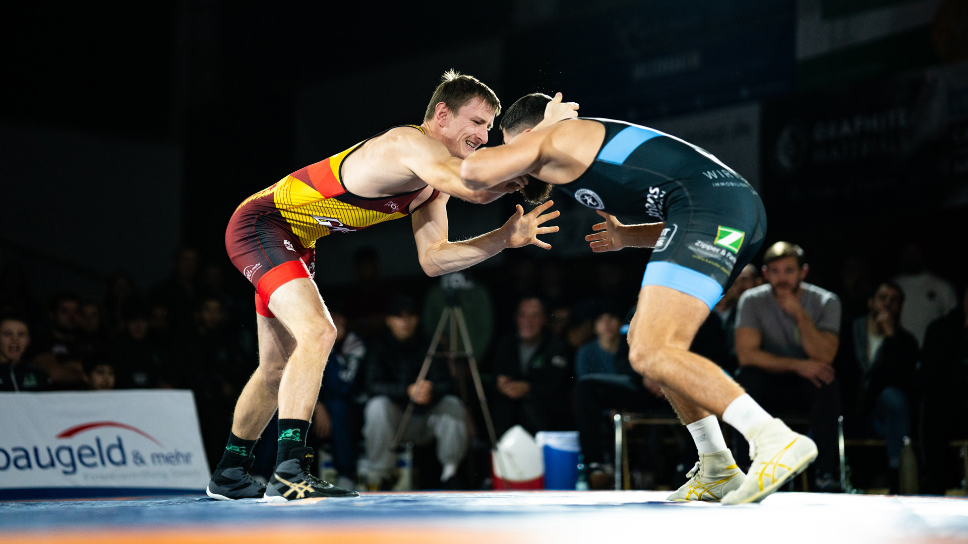 Two wrestlers face each other on a competition mat. One wears a red jersey.