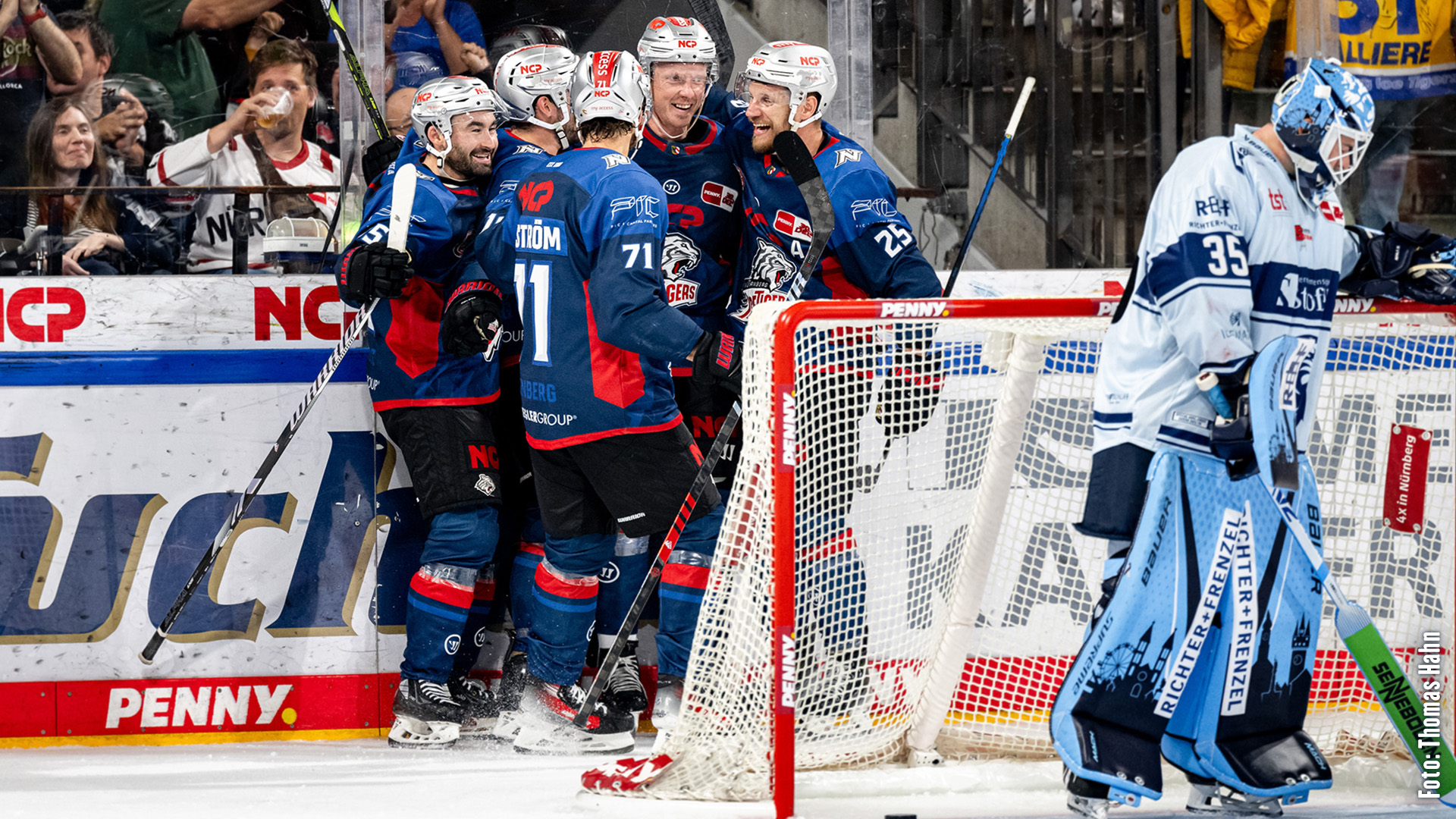 Four ice‑hockey players in blue jerseys celebrate a goal, while a goalkeeper stands nearby.