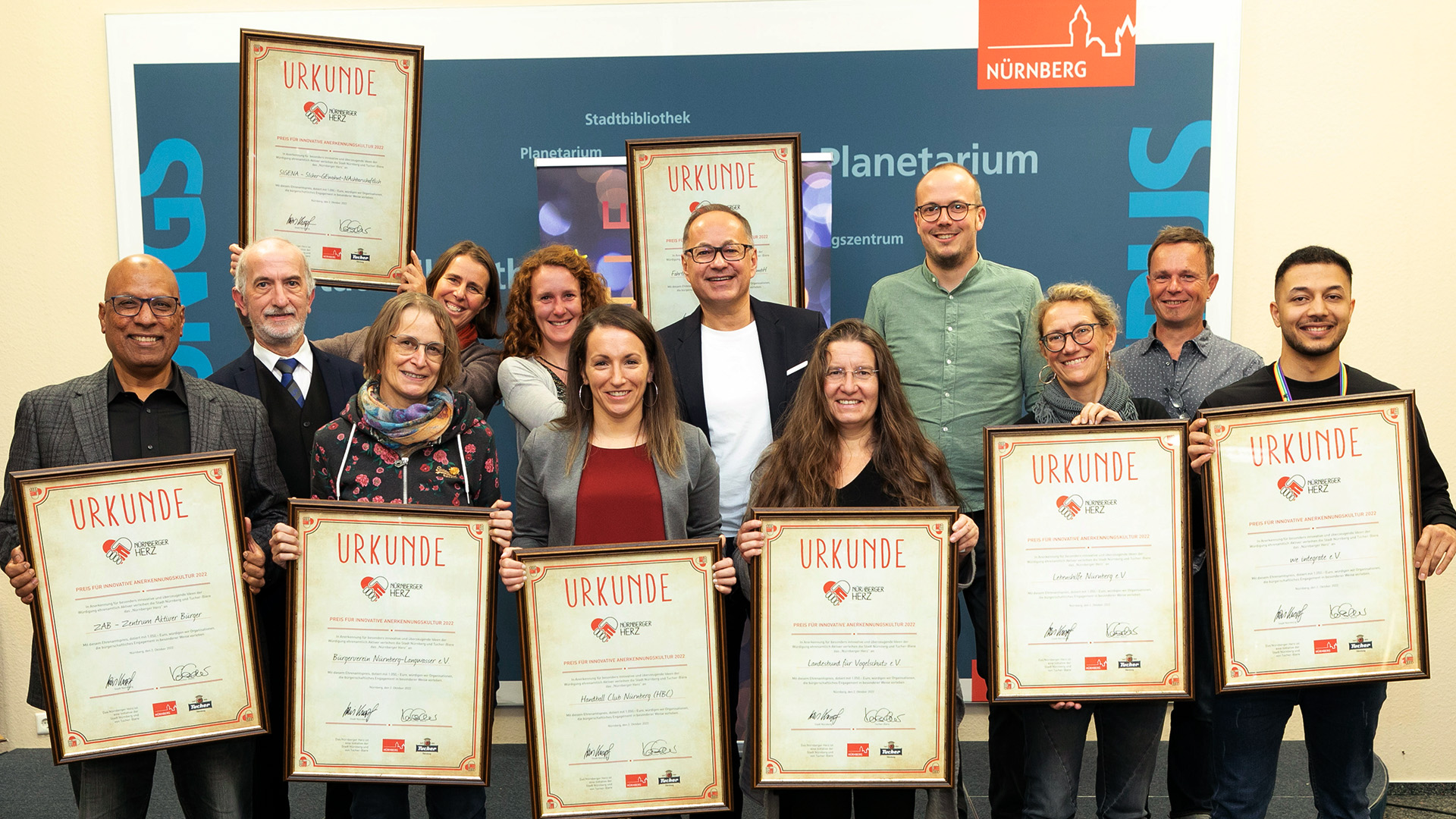 Eleven people stand side by side and proudly hold their certificates, they smile into the camera.