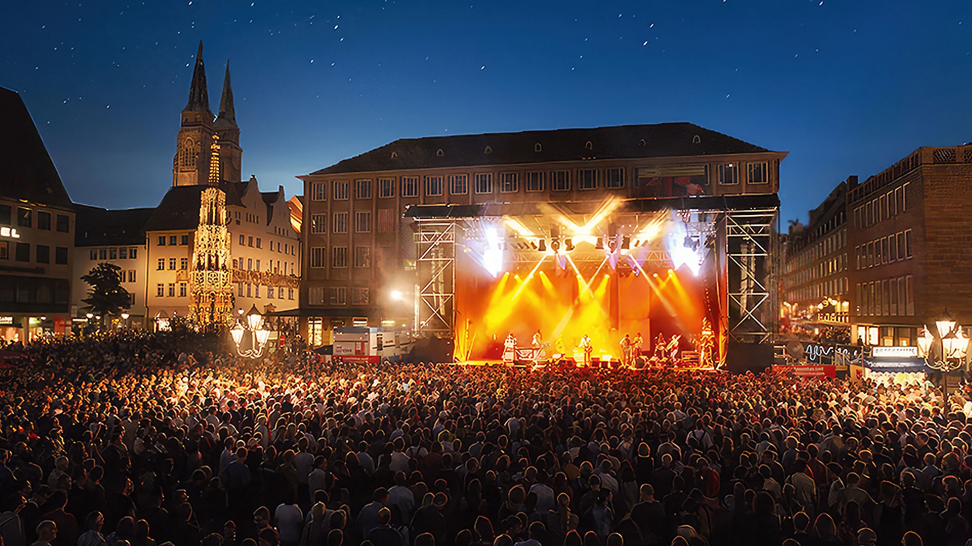 A lively open‑air night event on an illuminated square with numerous people.