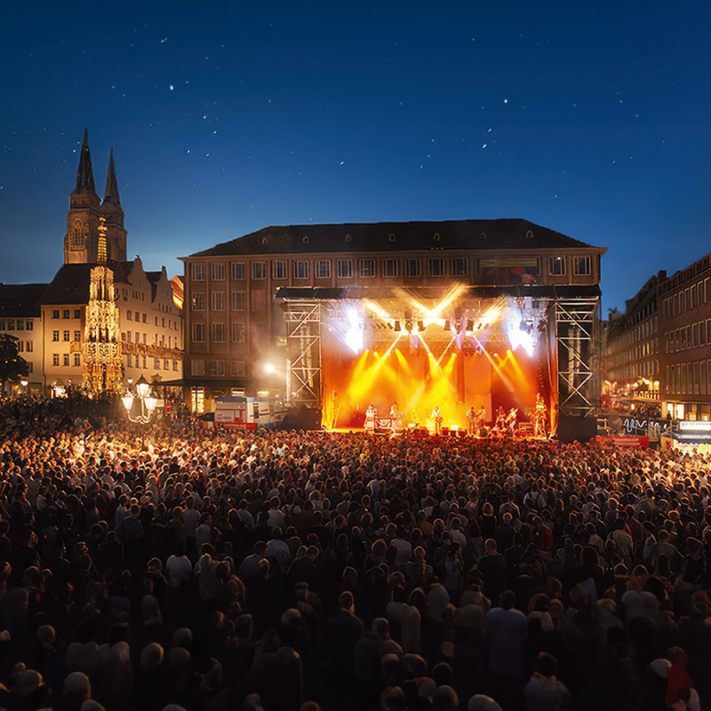 A large crowd stands in the evening on a square in front of a lit stage with musicians.